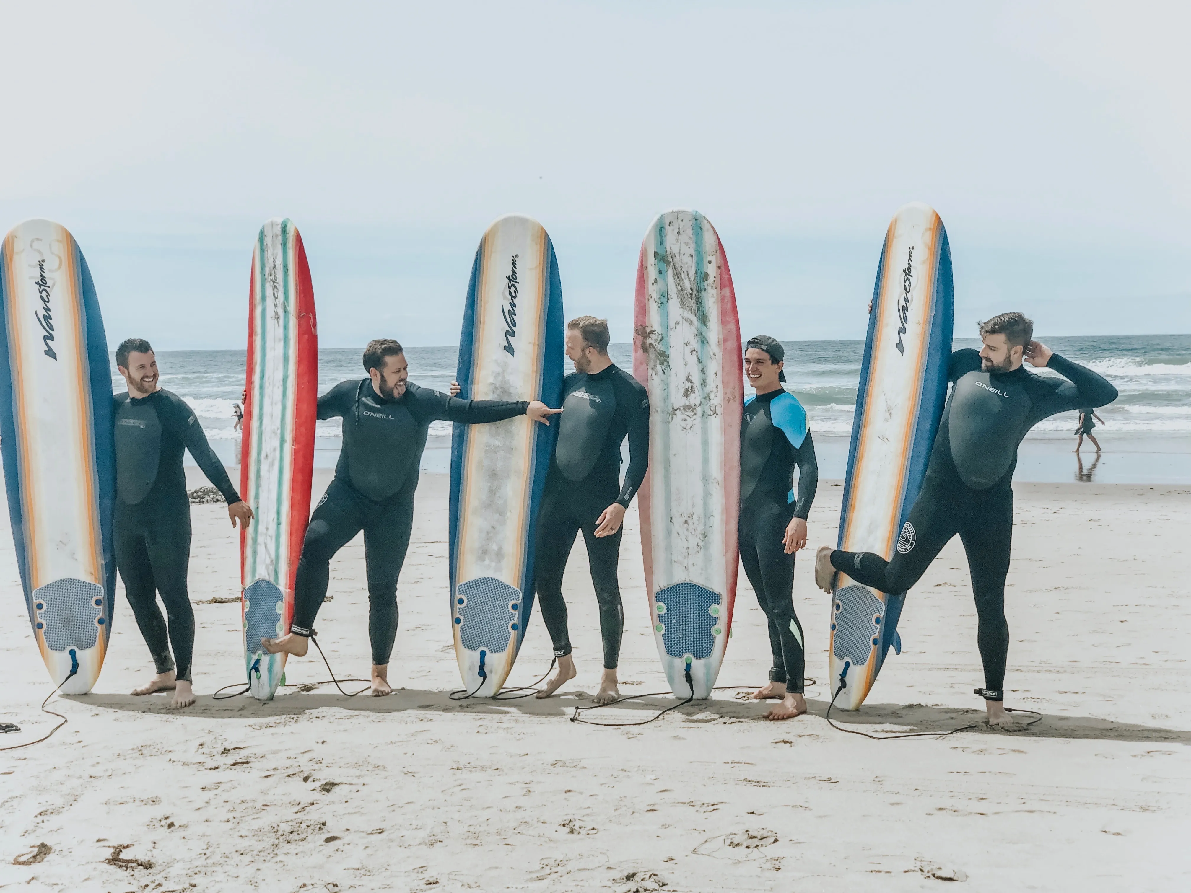 Six men in wetsuits posing with Wavestorm longboards on the beach